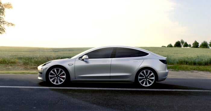 A silver Tesla Model 3 traveling on a road with a green field in background.