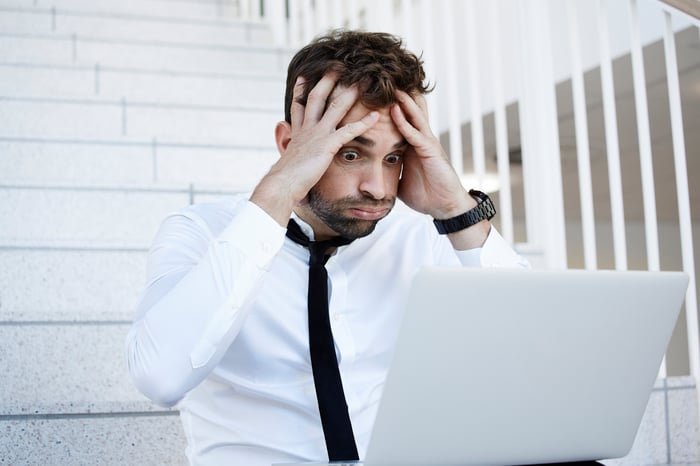 A visibly annoyed and worried young man grasping his head with both hands as he reads material on his laptop.