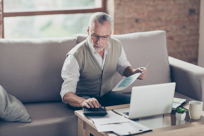 A senior man sitting on a sofa in front of a laptop holds a document in one hand while using a calculator.