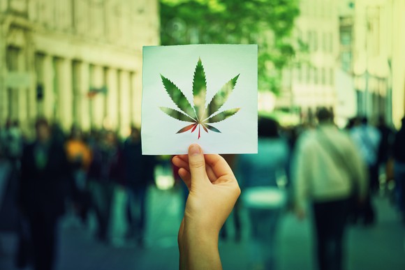 A hand holding up a piece of paper with a cutout shape of a marijuana leaf on it against a city street in the background.