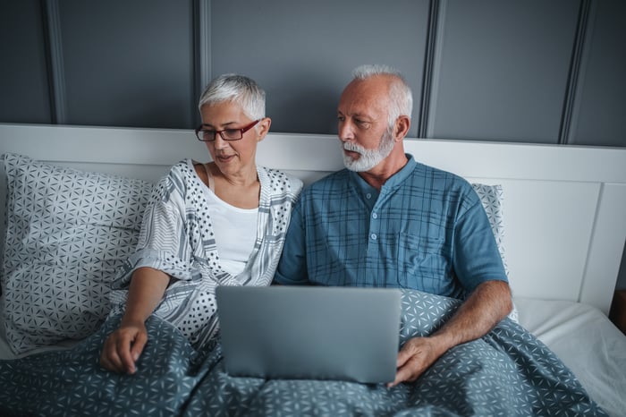 An older man and woman sitting up in bed, with the man holding an open laptop.