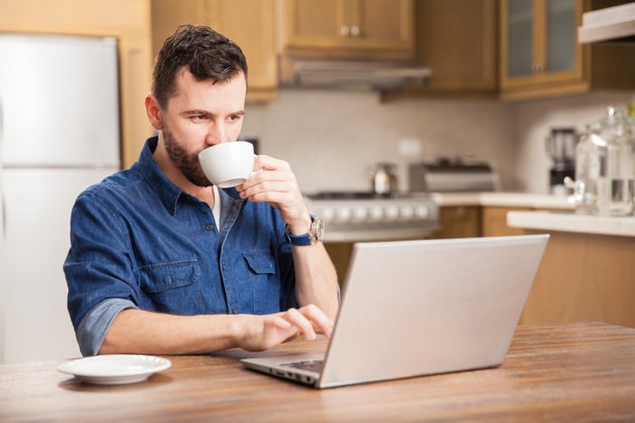 Man typing on laptop in kitchen while drinking from mug