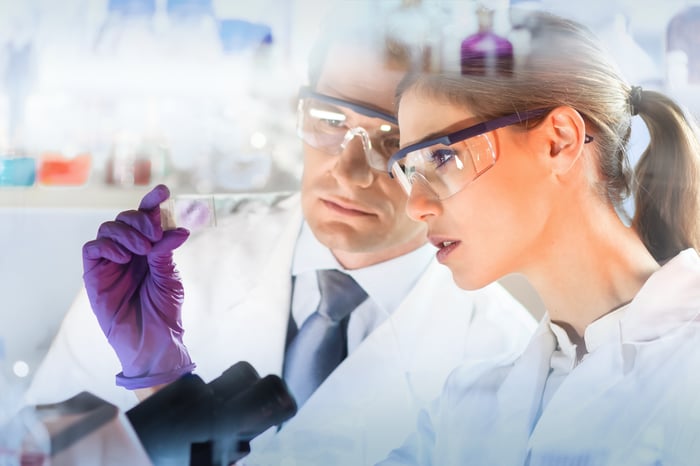 Two scientists in lab coats standing in front of a microscope and looking at a slide.