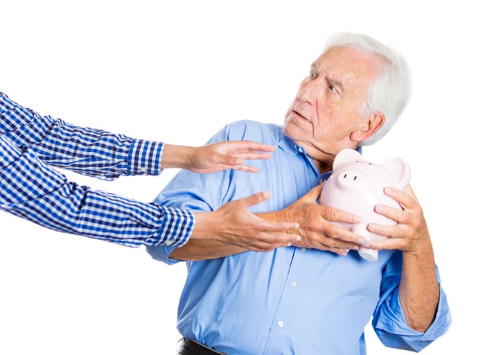 Older man holding piggy bank out of reach of grasping arms.