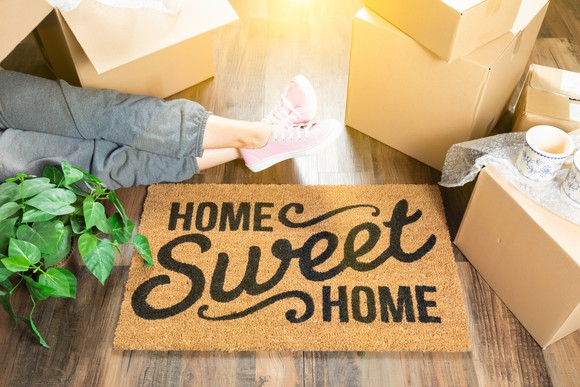 Woman sitting on floor next to plants, moving boxes, and mat that says home sweet home