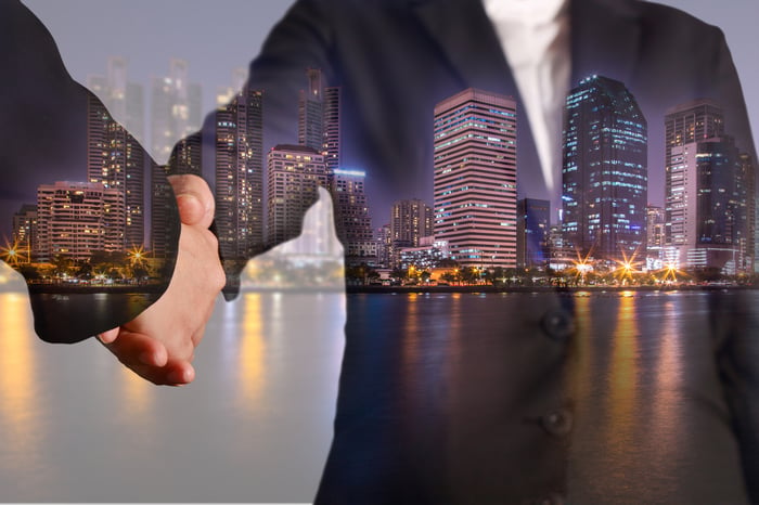 Two people shaking hands with a skyline of office building in the background.