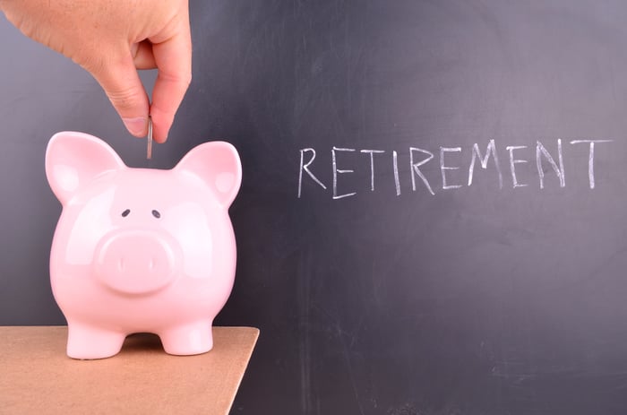 Coin being inserted into piggy bank that's sitting on a table in front of blackboard with the word retirement written in chalk