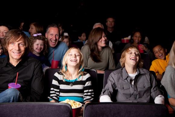 People of various ages -- some of whom are eating popcorn or drinking beverages -- sitting in seats in what appears to be a movie theater.