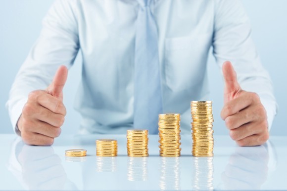 A businessman giving the thumbs-up sign with a growing stack of coins in front of him.