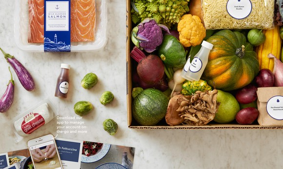 A Blue Apron meal kit, consisting of various ingredients neatly placed in a box and on a table.
