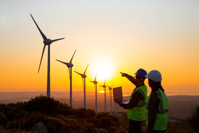 Two people in hard hats with a laptop looking at a row of wind turbines.