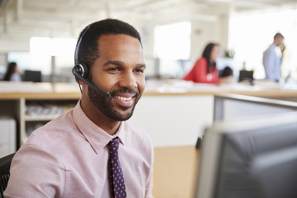 Smiling man wearing headset, and sitting in front of computer in an office.