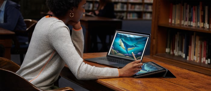 Woman using MacBook and iPad Pro at a desk in a library with bookshelves in the background