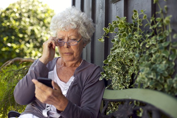 Senior woman sitting on a bench outdoors and adjusting glasses while looking at mobile phone