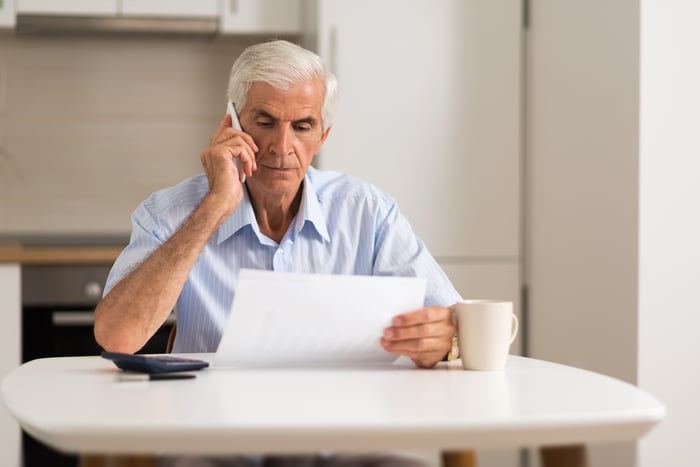 Senior man with a serious expression sitting at a kitchen table, holding document while talking on phone.