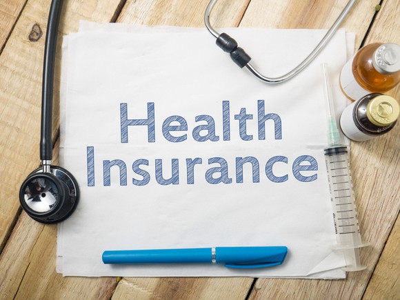 Piece of paper, with the words health insurance written on it, lying on wood surface with medical items (stethoscope, medicine) surrounding it.