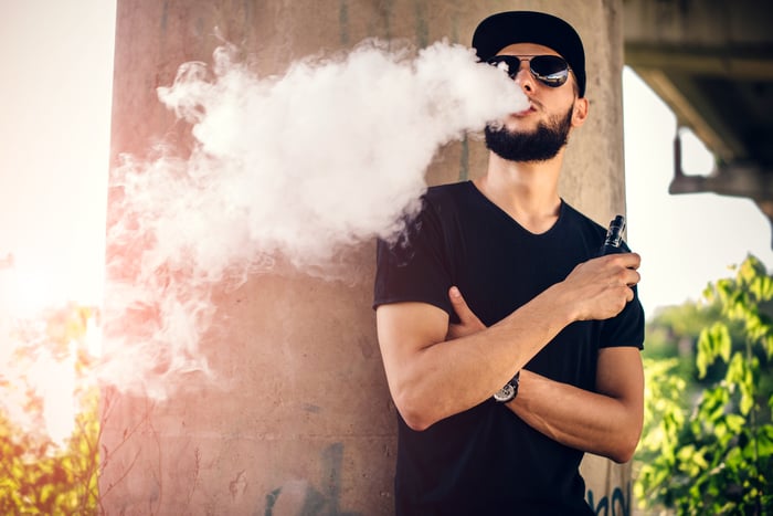 A young man with a beard blowing vape smoke from his mouth while outside.