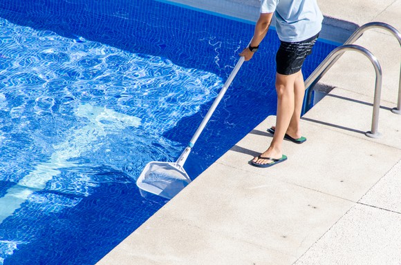 A man standing at the edge of a pool, skimming the surface with a cleaning net.