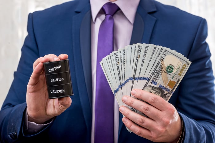 A man holding a tiny toy barrel of oil with "caution" written on it in one hand and cash in the other hand.