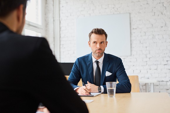 Man in suit with serious expression jotting down notes while seated across from another man