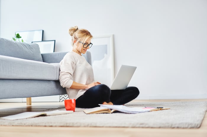 Woman sitting on floor up against couch, typing on laptop.