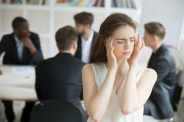 Woman sitting in front of a table of men in suits, covering her ears