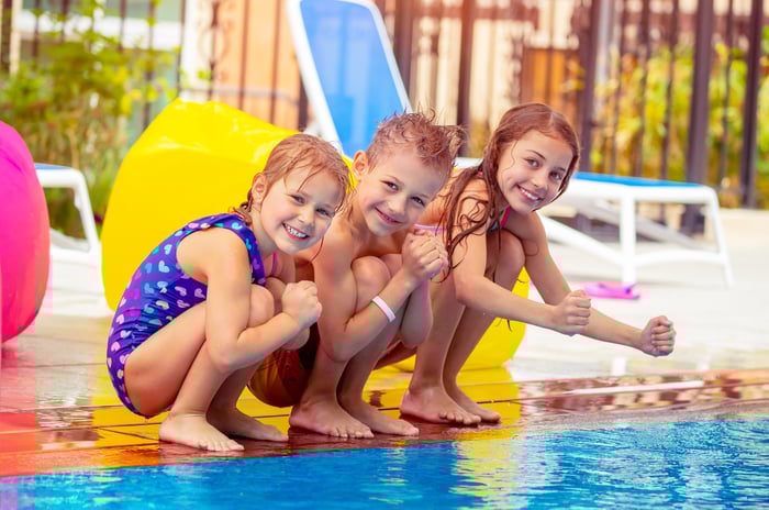 Three smiling young children perched on the edge of a swimming pool