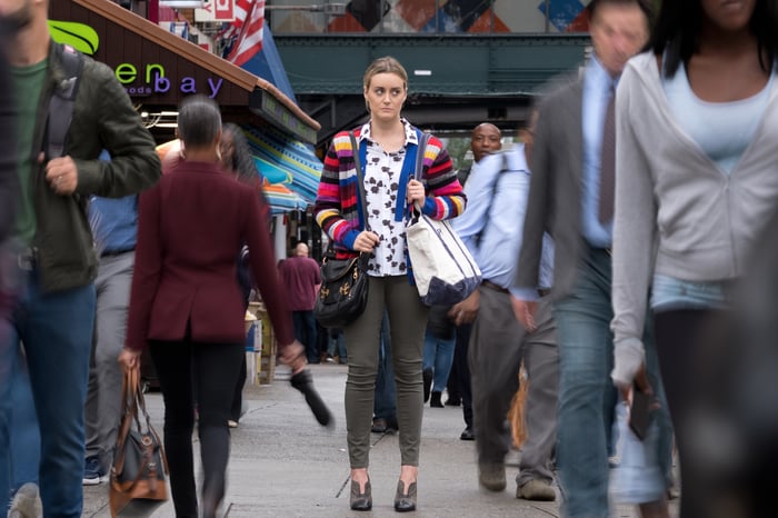 A woman standing in a crowded street as throngs of pedestrians pass her on both sides.