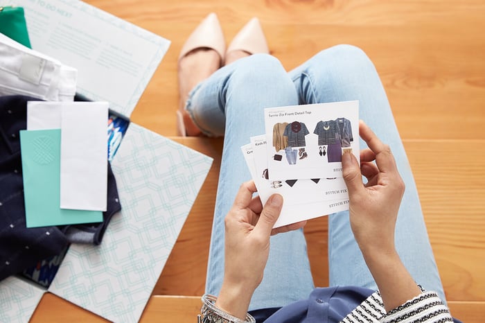 A woman holding style cards from Stitch Fix with an open box full of new clothes laying on the floor.