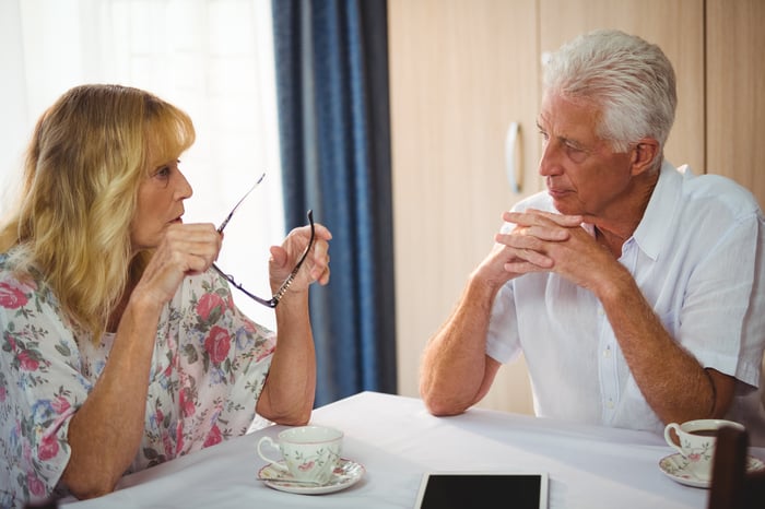 Older man and woman sitting at a table with teacups in front of them, sporting serious expressions.