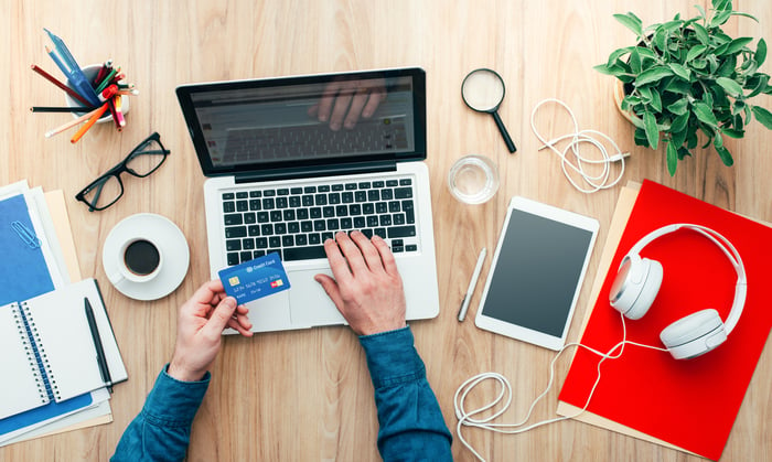 A man makes a credit card purchase using his laptop at his desk, which is surrounded by glasses, a cup of espresso, a note pad, headphones, a magnifying glass, a plant, and a tablet.