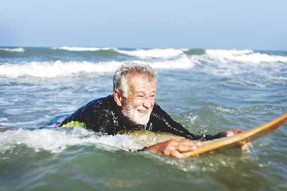 An older senior rides on his stomach on a surfboard in the ocean.
