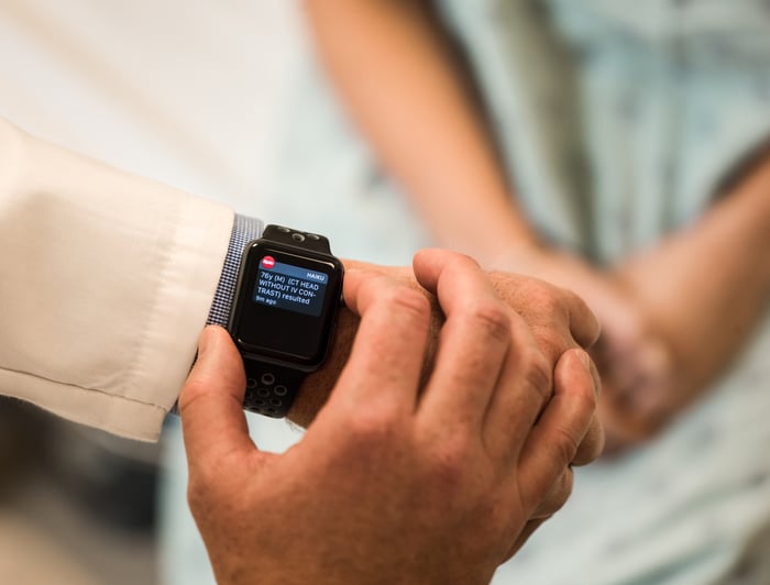 A physician reviewing an Apple Watch notification as a patient sits on a bed nearby.