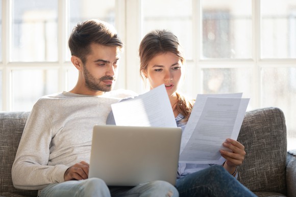 Somber-looking man and woman using laptop on couch and holding sheets of paper.
