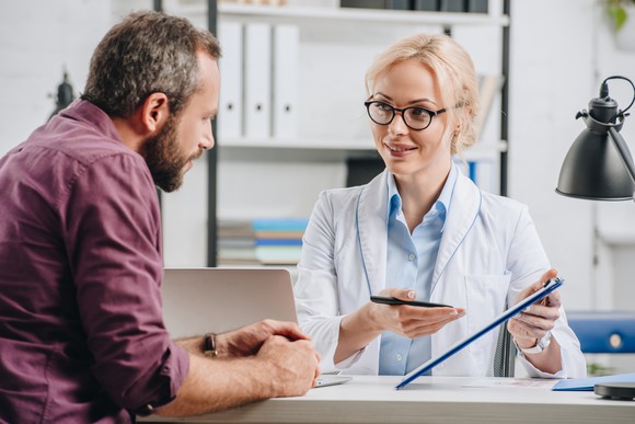 Woman in white coat gesturing toward clipboard while man looks on