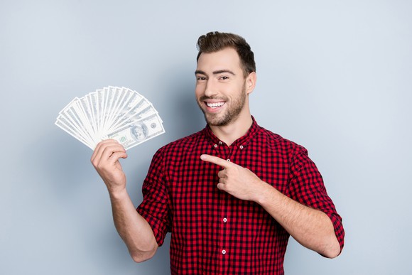 Smiling man fanning out a bunch of hundred-dollar bills and pointing toward them.