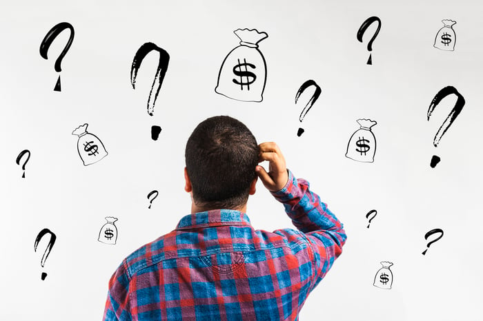 Man with hand on head is facing a wall with question marks and drawings of money bags.