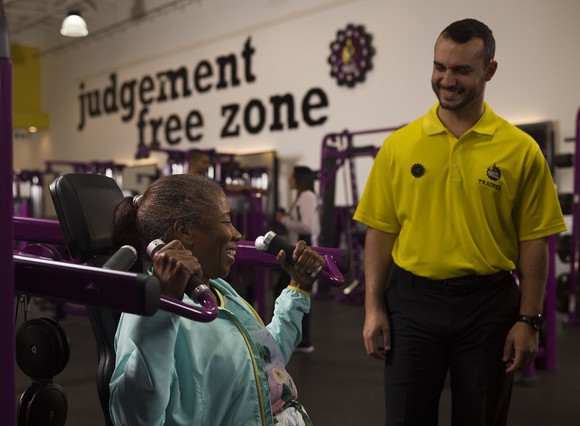 A Planet Fitness trainer smiling at a woman exercising.