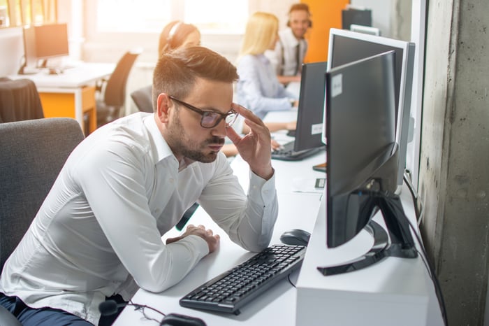 Man with sour expression sitting at computer, adjusting eyeglasses