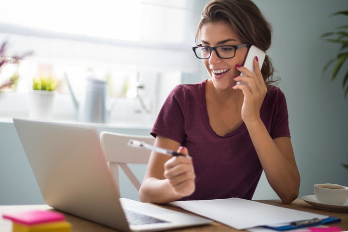 Woman holding pen and talking on phone while sitting at laptop