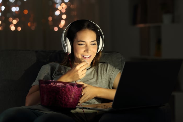 Woman wearing headphones and eating popcorn while watching streaming video on a laptop in a dark room.