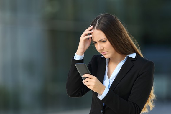 A young woman in a business suit holding her phone in one hand and frowning.