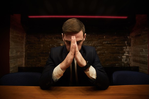 A man sitting at a table with his head bowed and his hands palm to palm in front of his face.