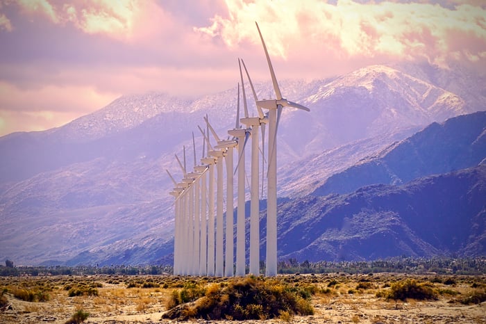 A line of wind turbines with mountains in the background.