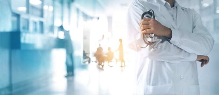 Man in white coat in hospital hallway with arms crossed while holding a stethoscope