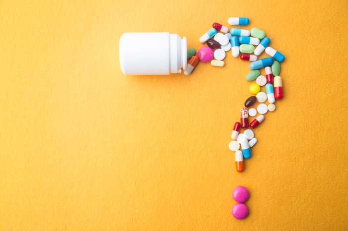 A white pill bottle with various pills and tablets spilling out it, forming a question mark.