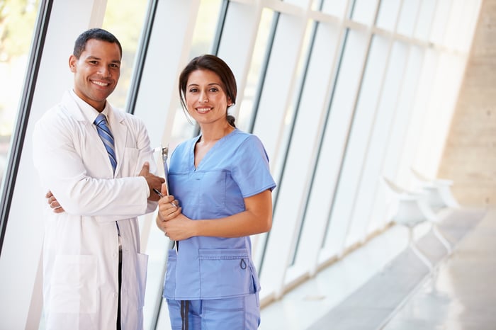 Male physician in white coat standing next to female healthcare provider in blue scrubs in a hospital hallway, both smiling.
