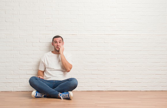 Man in jeans and t-shirt sitting against a white brick wall, biting his nails