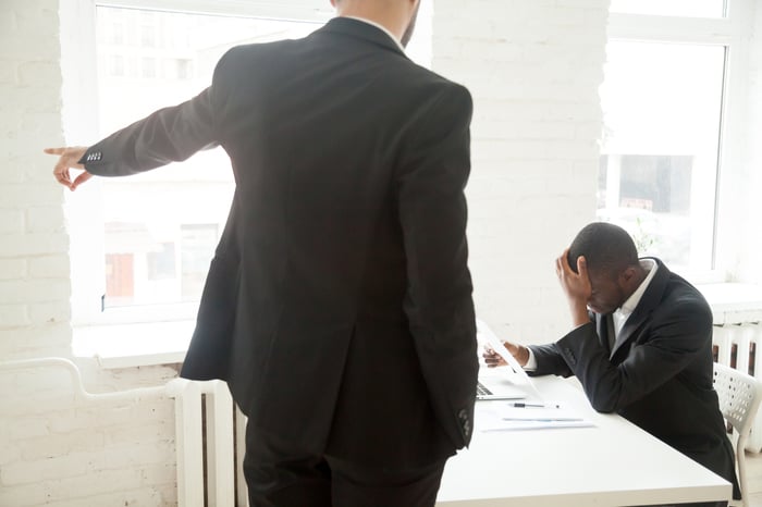 Man in suit standing and pointing, while another man is seated and holding his head in his hand.