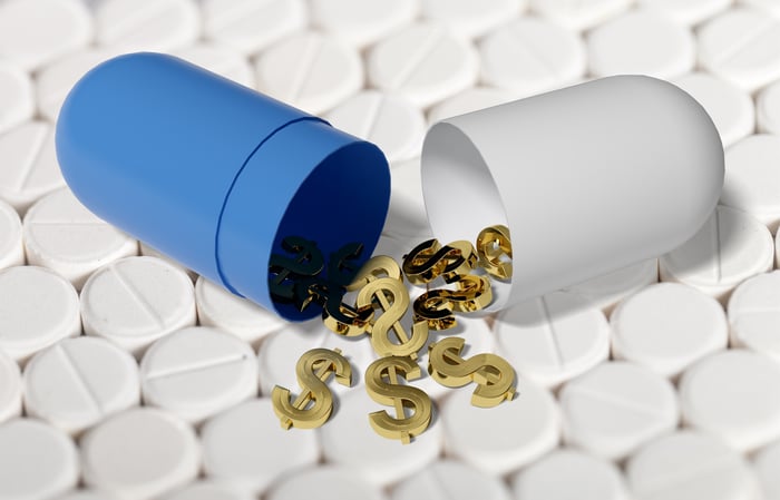 Split apart pill capsule on top of  small white tablets with golden dollar signs coming out of the capsule